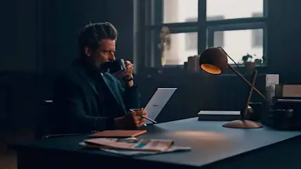 Man in a dark suit drinks from a cup, seated at a desk with a lamp, laptop, notepad, and newspaper near a window.