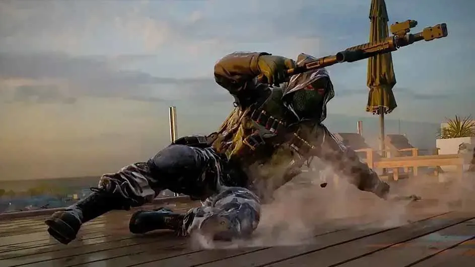 Een persoon in camouflage kleding en een bivakmuts ligt op een houten vloer en houdt een geweer vast. Op de achtergrond een strand.