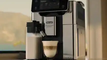 De’Longhi advertentie met een zilveren volautomatische espressomachine die een cappuccino zet in een moderne keuken met berguitzicht.