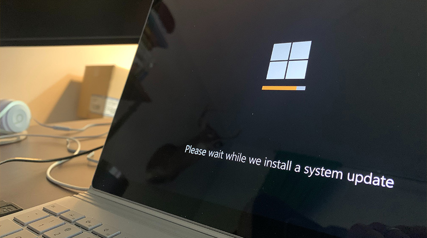 Laptop scherm toont "Please wait while we install a system update" met Windows logo en oranje voortgangsbalk.