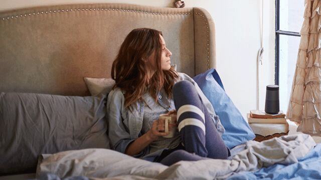 Vrouw zit in bed met een kopje. Kussen, beddengoed en een luidspreker staan op het nachtkastje naast het raam.