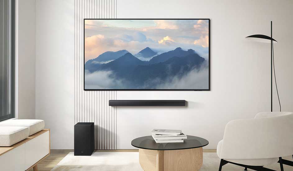 Een moderne woonkamer met een grote tv aan de muur, een zwarte soundbar eronder, een subwoofer op de vloer en een ronde salontafel.