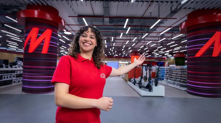 Medewerker in rood poloshirt wijst uitnodigend naar de ingang van een elektronicawinkel met fel verlichte displays en grote zuilen met een M-logo.