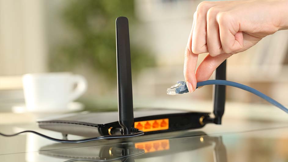 Een hand steekt een blauwe netwerkkabel in een zwarte router met oranje poorten. Op de achtergrond staat een witte mok op tafel.