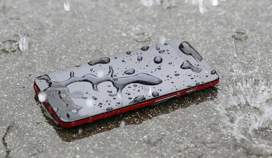 A red smartphone lies face up on wet pavement, its dark screen covered in raindrops as splashes ripple across the surrounding puddle.