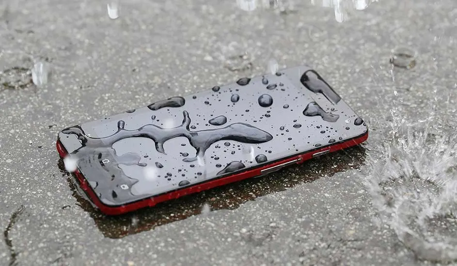 A red smartphone lies face up on wet pavement, its dark screen covered in raindrops as splashes ripple across the surrounding puddle.