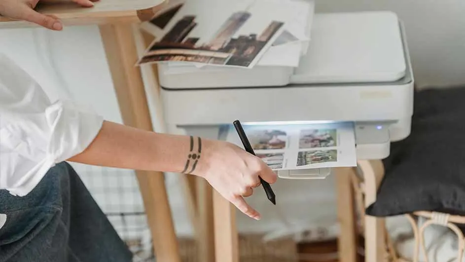 Een persoon haalt een afdruk uit een witte printer op een houten tafel. Er liggen foto's bovenop de printer.