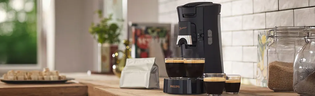 Op het aanrecht staat een Philips Senseo-koffiemachine met drie kopjes koffie, omringd door potten en een zak koffie.