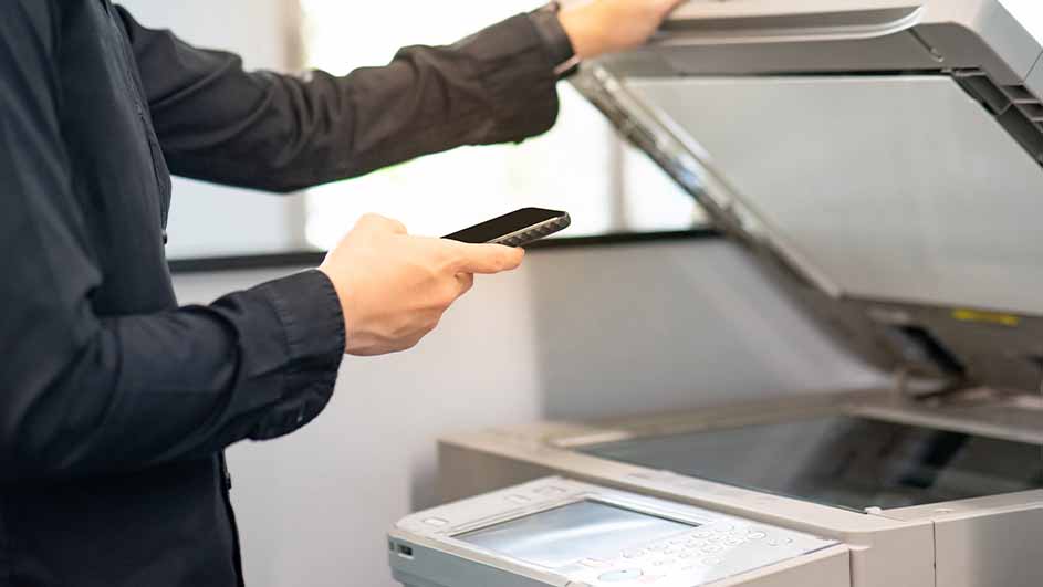Een persoon in een zwart overhemd bedient een grijze printer en houdt een zwarte telefoon vast.
