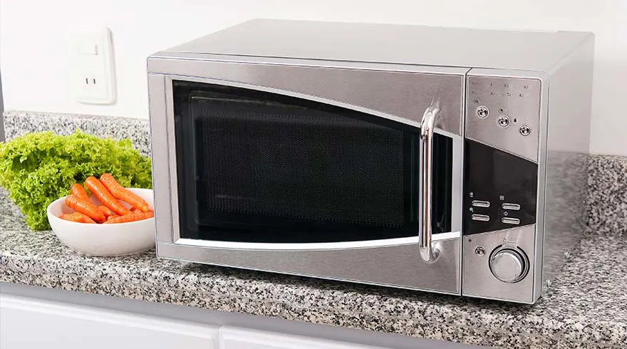 Een zilverkleurige magnetron staat op een aanrecht naast een witte kom met wortelen en een krop sla.