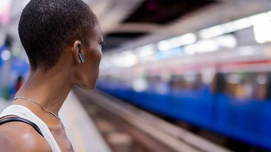 A person with an earpiece waits on a platform beside train tracks. A blue train is out of focus in the background.
