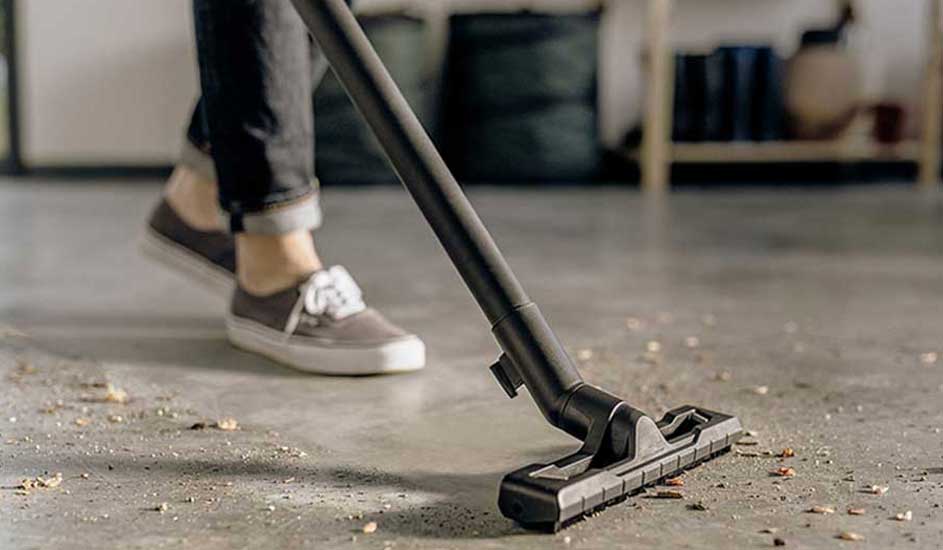 Iemand stofzuigt een betonnen vloer vol kruimels; de zwarte zuigmond schuift voorwaarts terwijl sneakers en opgerolde jeans in beeld zijn.