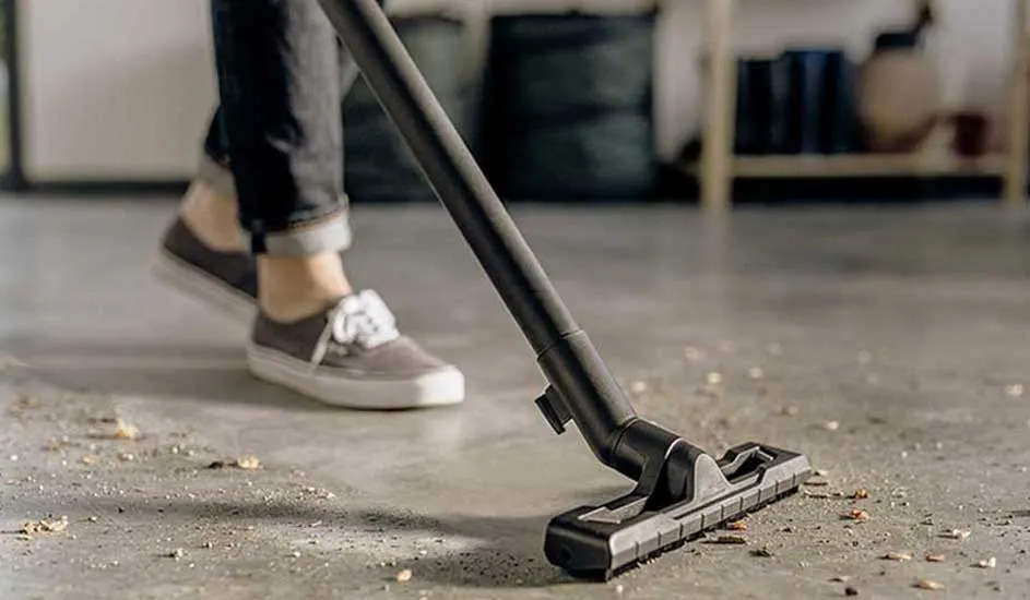 Iemand stofzuigt een betonnen vloer vol kruimels; de zwarte zuigmond schuift voorwaarts terwijl sneakers en opgerolde jeans in beeld zijn.