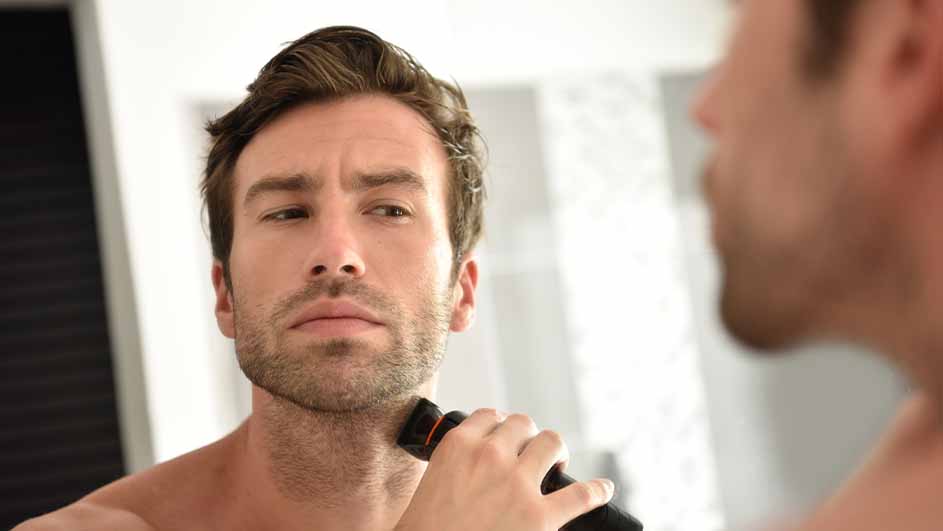 A man shaves his face with an electric razor in front of a mirror.