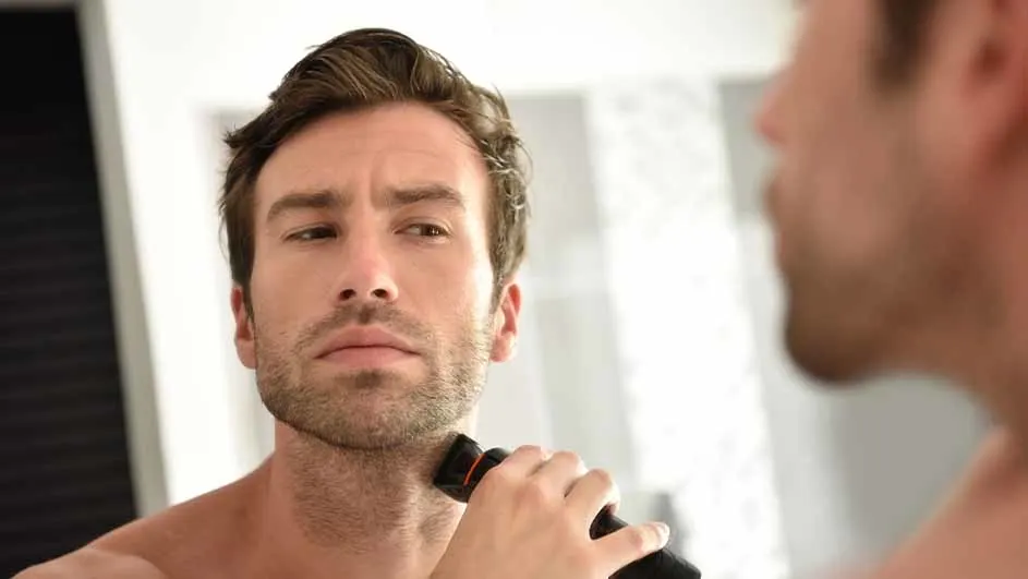 A man shaves his face with an electric razor in front of a mirror.