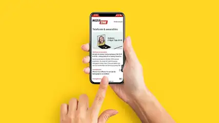 Hands hold a smartphone displaying a "Media Zine" article on a yellow background. A finger touches the screen.