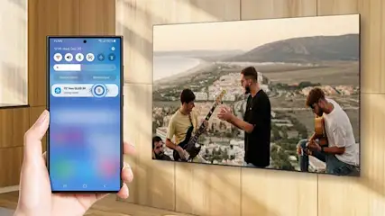 Smartphone met smart home app bedient Samsung Neo QLED 8K TV waarop muziekband speelt tegen mediterraan berglandschap in houten woonkamer.