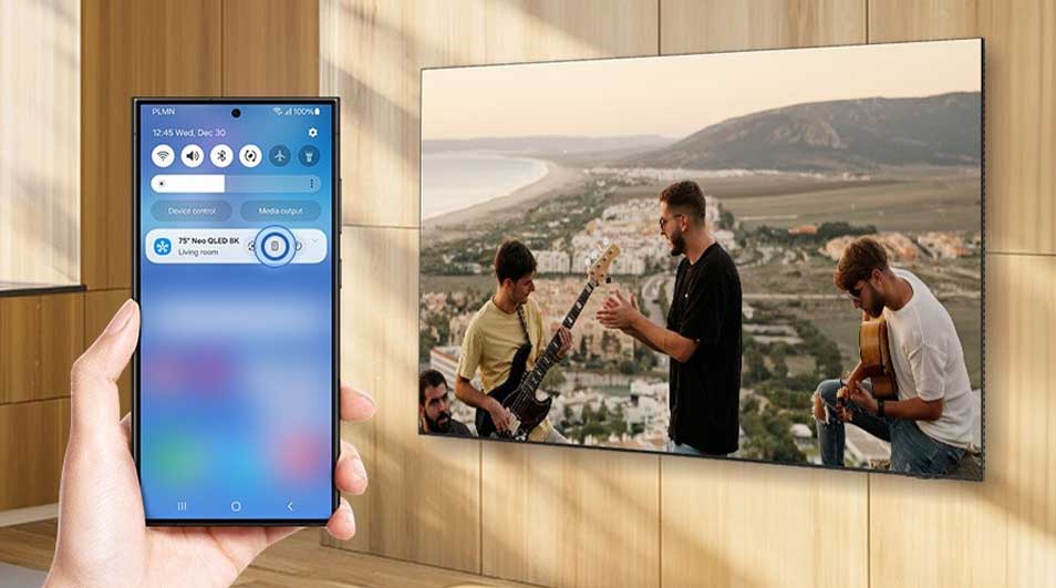 Smartphone with smart home app controlling Samsung Neo QLED 8K telly showing music band against Mediterranean mountain landscape backdrop.