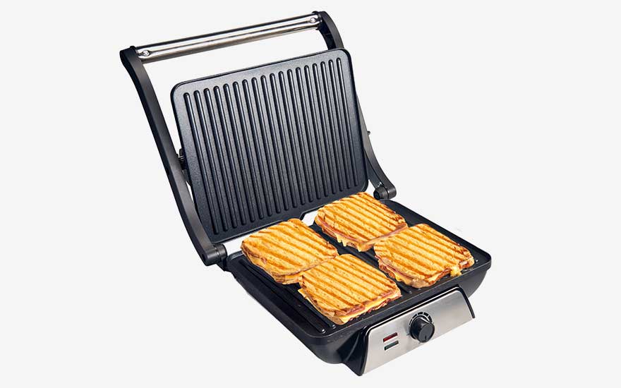 Large contact grill opened flat, holding four golden paninis with clear grill lines, metal handle and front dial to adjust the heat.