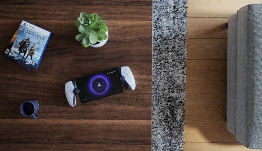 Op een houten tafel liggen een spelletje, een plant, een apparaat en een mok, naast een grijze mat en een bank.