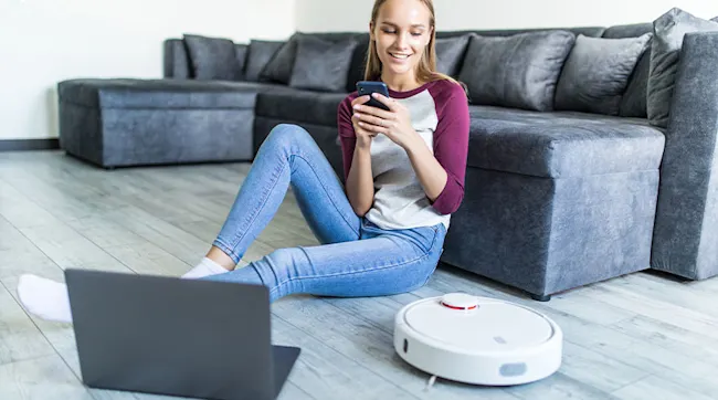 Vrouw zit op de vloer met een telefoon. Er is een grijze bank, een laptop en een robotstofzuiger te zien.