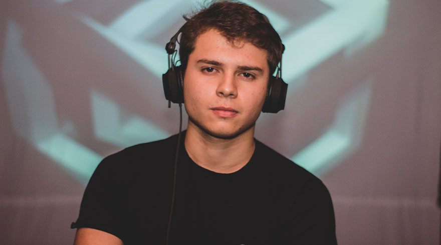 Een persoon met een zwarte T-shirt en een koptelefoon staat voor een muur met een lichtgevend grafisch patroon.