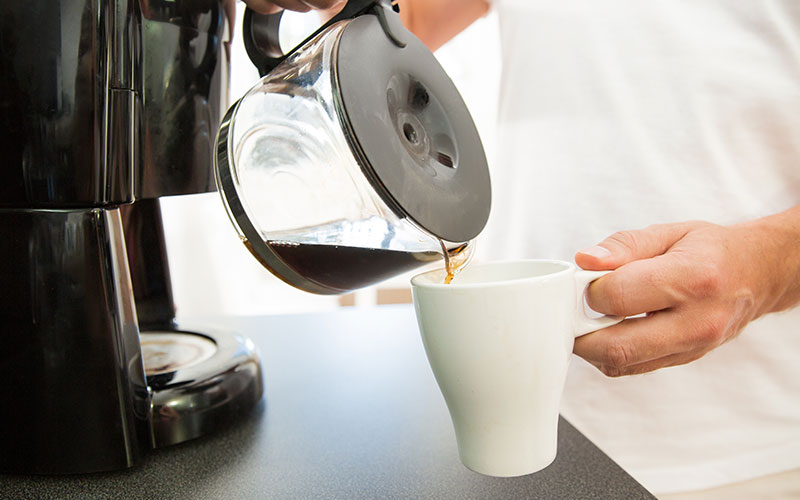 Hand schenkt vers gezette koffie uit een glazen kan in een witte mok naast een koffiezetapparaat.