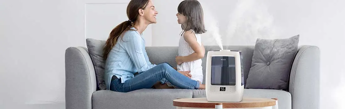 Een moeder en dochter zitten samen op een bank, glimlachen naar elkaar naast een luchtbevochtiger die stoom afgeeft op de salontafel.