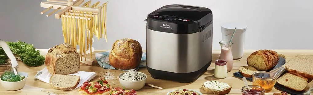A Tefal bread maker sits on a wooden table, surrounded by fresh loaves, spreads, yoghurt and jars of jam for a generous meal.