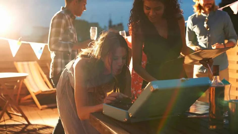 Mensen op een terras met een platenspeler, drankjes, een tafel en stoelen. Banners hangen op de achtergrond.
