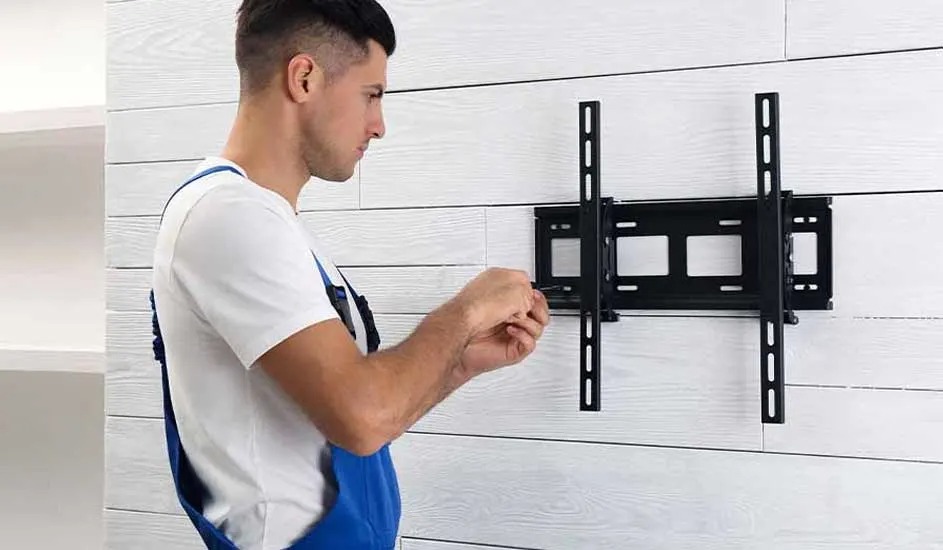 A technician in blue overalls is fitting a black TV wall bracket to a white wooden wall in a modern, bright room
