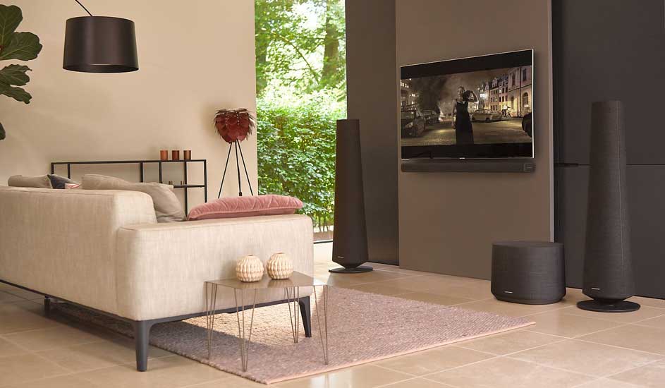 Stylish living room with a grey sofa, glass table, and soundbar setup. Wall-mounted TV playing a film, with a green view through windows.