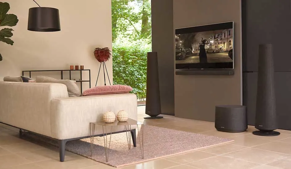 Stylish living room with a grey sofa, glass table, and soundbar setup. Wall-mounted TV playing a film, with a green view through windows.