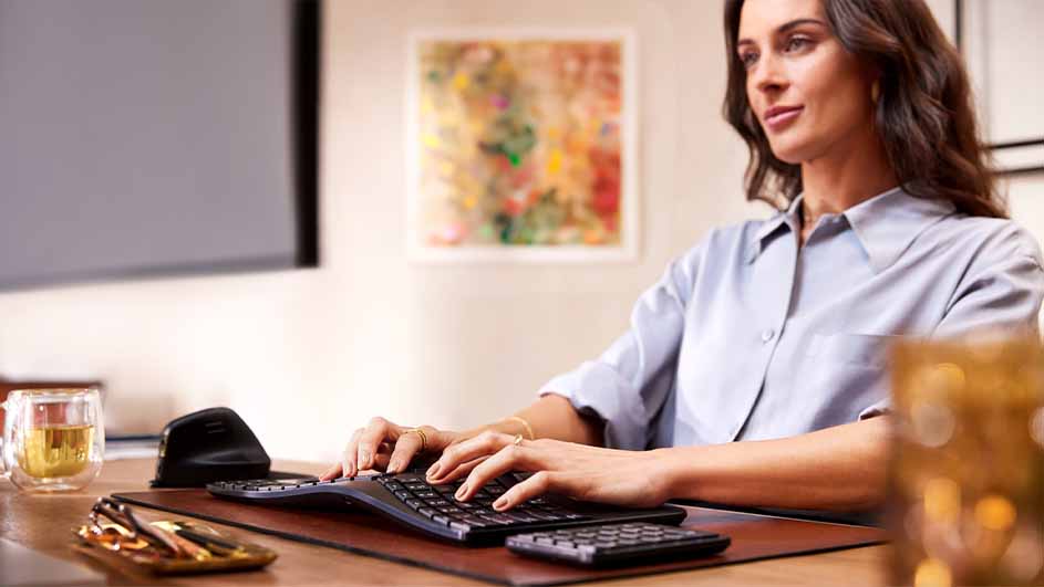 Vrouw typt op zwart toetsenbord op een bruin bureau. Een mok, muis en pennen liggen naast het toetsenbord. Aan de muur hangt een schilderij.