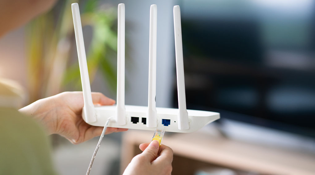 Iemand sluit een gele netwerkkabel aan op een witte router met vier antennes. Op de achtergrond staat een tv.