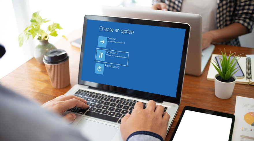 Een persoon typt op een laptop met een blauw scherm met opties. Op tafel staat een tablet, koffie, een plant en papieren.