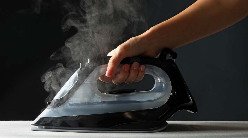 Een hand houdt een stoomstrijkijzer vast dat stoom afgeeft op een gestreepte ondergrond, tegen een donkere achtergrond.