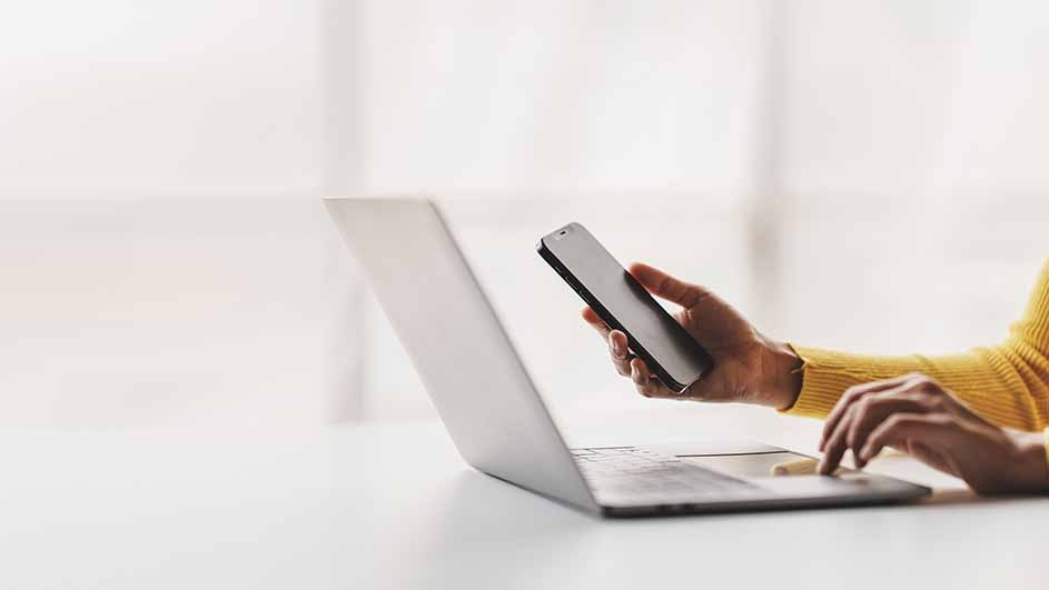 A person types on a laptop while holding a smartphone. A bright window is in the background.