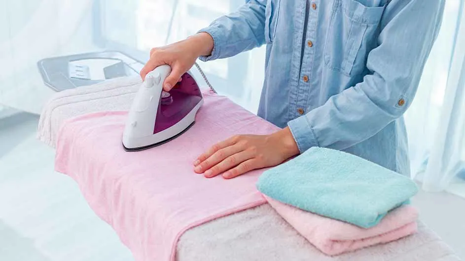 Een persoon strijkt een roze doek met een wit en paars strijkijzer op een strijkplank, met opgevouwen doeken ernaast.
