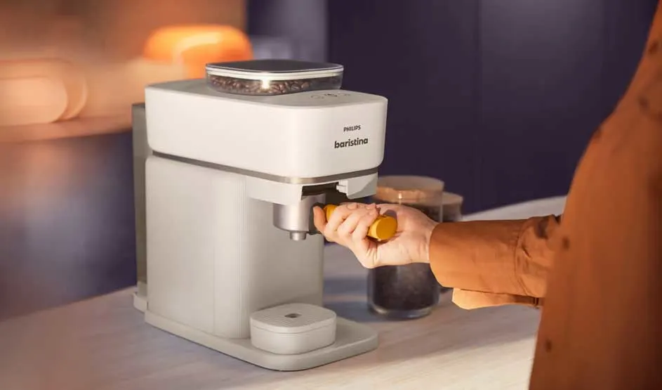 Een persoon plaatst een portafilter in een witte koffiemachine, met een glas koffie ernaast en warme verlichting op het aanrecht.
