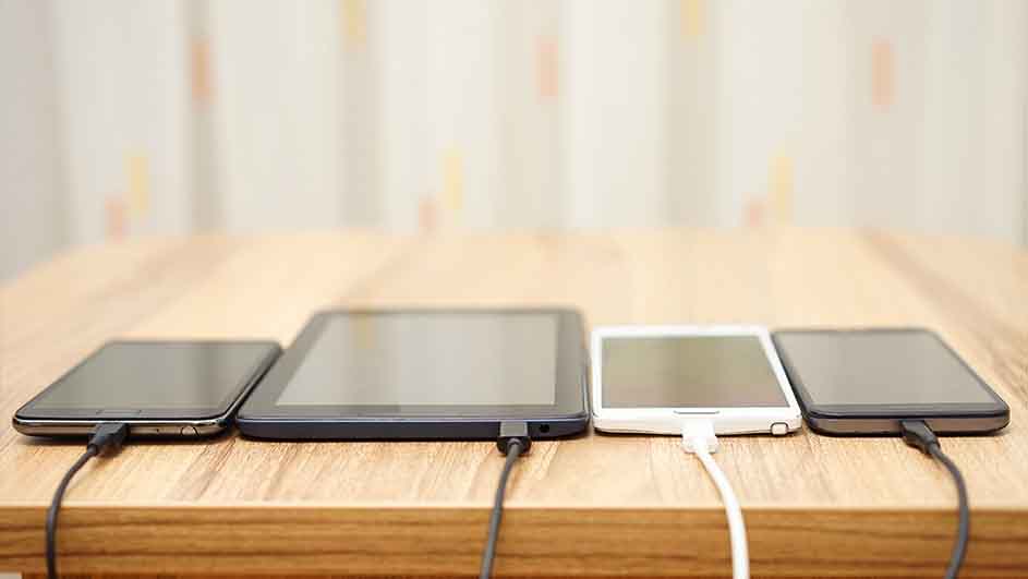 Vier telefoons en een tablet liggen op een houten tafel en laden op met zwarte en witte kabels.