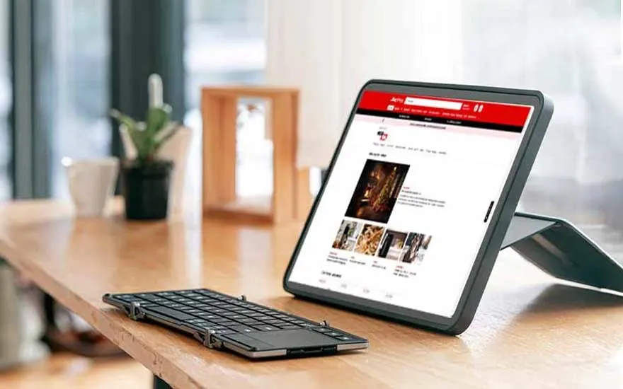 On a wooden desk a tablet on a stand shows an online shop page, placed beside a folding keyboard and a simple white coffee mug.