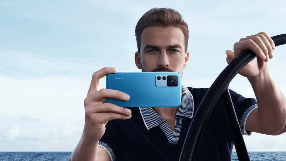 Een man houdt een blauwe telefoon horizontaal vast met de camera naar voren gericht, tegen een achtergrond van zee en lucht.