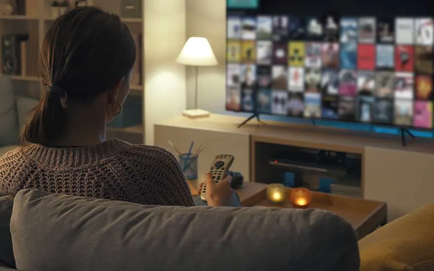 A woman sits on a sofa in a cosy room holding a remote, ready to browse and watch content on a large television screen.
