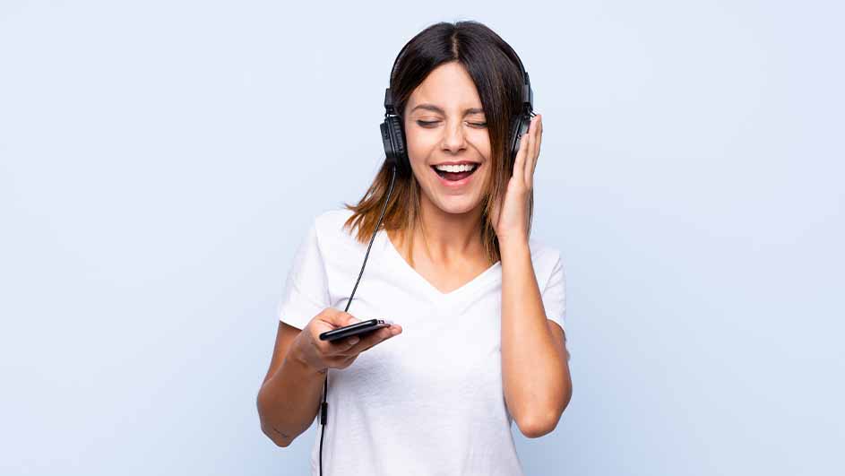 Een vrouw lacht en luistert naar een koptelefoon, terwijl ze een smartphone in haar hand houdt, tegen een blauwe achtergrond.