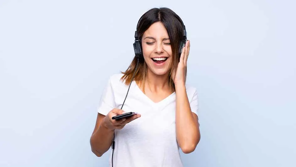 Een vrouw lacht en luistert naar een koptelefoon, terwijl ze een smartphone in haar hand houdt, tegen een blauwe achtergrond.