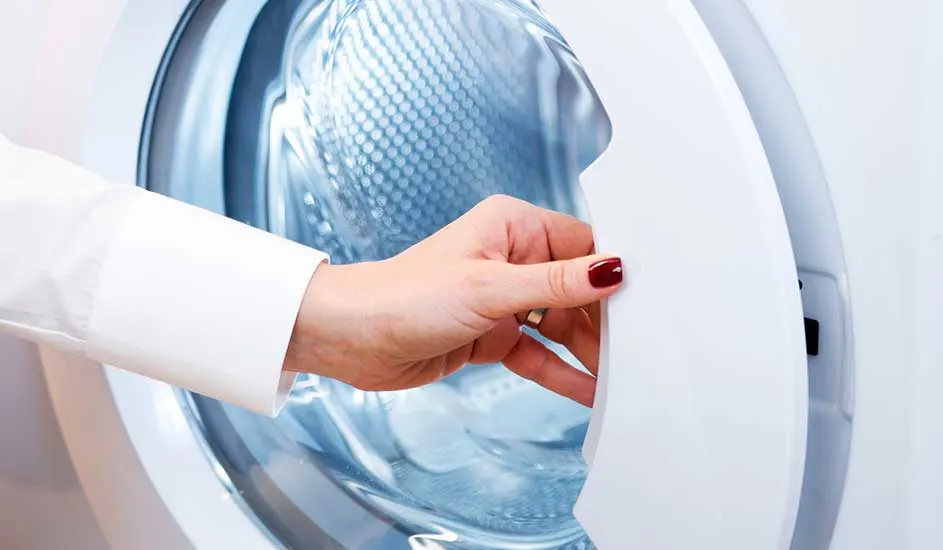 Een hand in een witte blouse opent de witte deur van een wasmachine met een blauwe trommel.