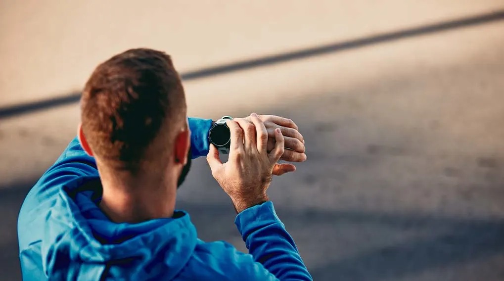 A man in a blue hoodie checks a black sports watch on his wrist outdoors.