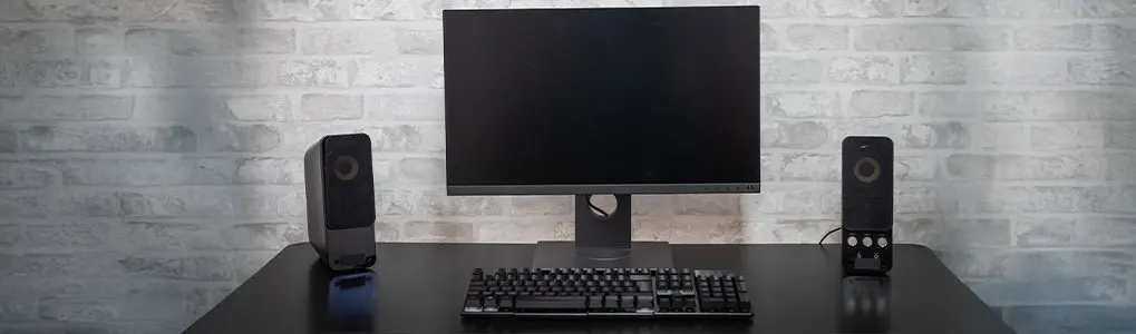 A desk has a monitor, keyboard, and two speakers. The background is a brick wall.