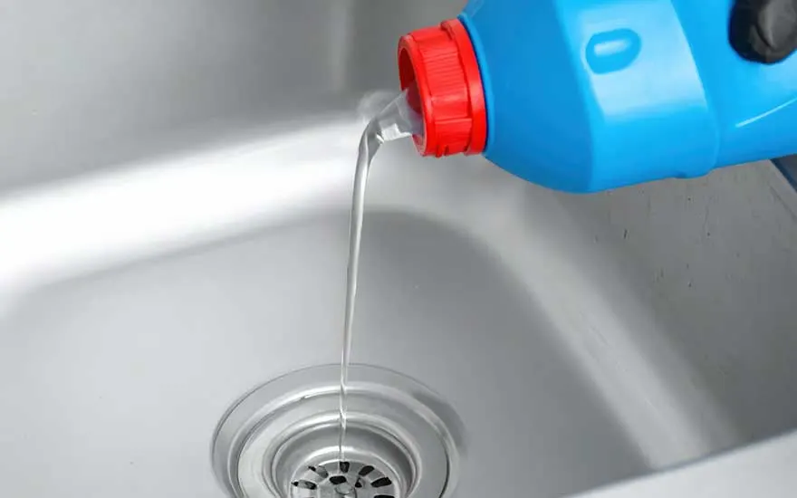 Een blauwe plastic fles met rode dop waaruit een heldere vloeistof wordt gegoten in een witte gootsteen. De vloeistof stroomt direct richting de metalen afvoer. Vermoedelijk is dit een vloeibaar ontstoppingsmiddel dat wordt gebruikt om een verstopping te verhelpen.